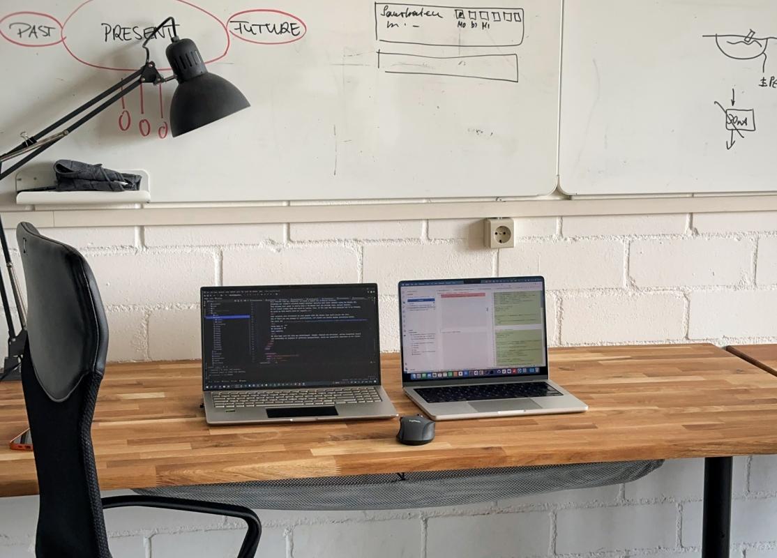 Moderner Arbeitsplatz mit zwei Laptops, einer Maus und einem Bürostuhl vor einem Whiteboard, auf dem verschiedene Diagramme und Notizen zu sehen sind.