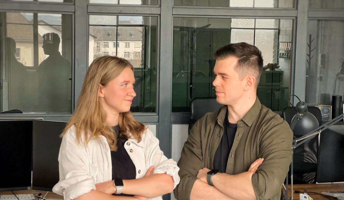 Zwei junge Menschen stehen sich im Büro gegenüber, mit verschränkten Armen und einem selbstbewussten, freundlichen Blickkontakt.