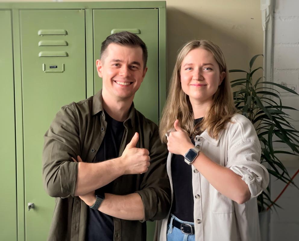 Ein Mann und eine Frau stehen lächelnd vor einem grünen Schrank und zeigen mit erhobenen Daumen eine positive und motivierte Haltung.