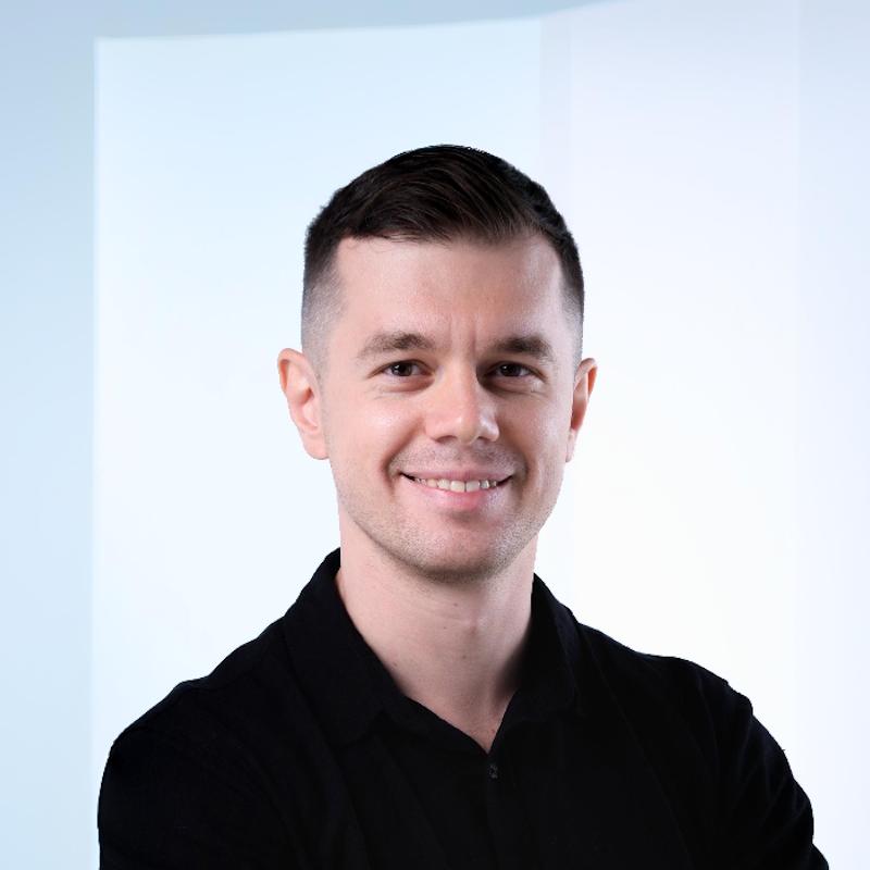 Junger Mann sitzt entspannt und lächelnd in einem modernen Büro mit grünen Pflanzen im Hintergrund.