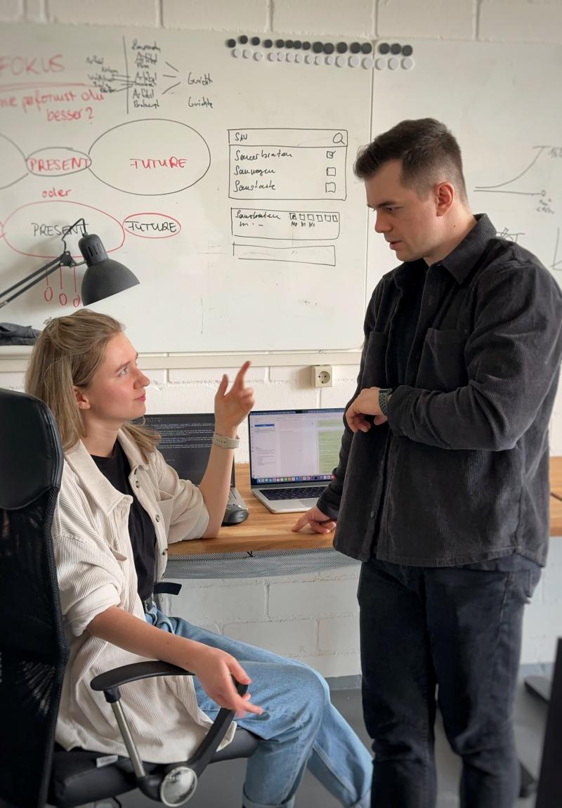 Zwei Kollegen diskutieren in einem Büro mit Whiteboard und mehreren Laptops; die Frau sitzt am Schreibtisch und erläutert etwas, während der Mann stehend zuhört und auf seine Smartwatch blickt.